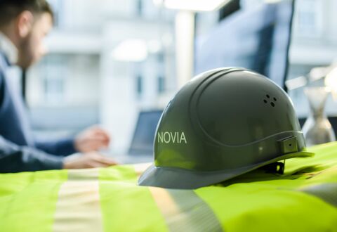 NOVIA GmbH Consulting und Baumanagement - Detailaufnahme: Gelbe Warnweste und Baumhelm mit NOVIA-Logo liegen auf Schreibtisch eines Mitarbeiters, der im Hintergrund zu sehen ist.